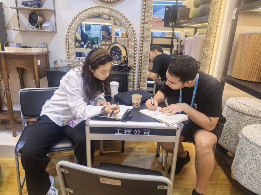 Trade show visitors reviewing notes together beside decorative mirror and upholstered furniture samples at the Canton Fair.